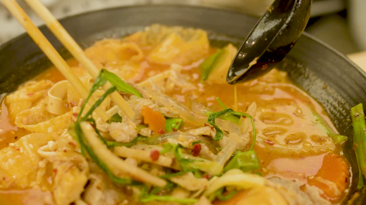 Chopsticks and spoon lift ingredients from spicy Asian noodle soup in warm, well-lit kitchen setting