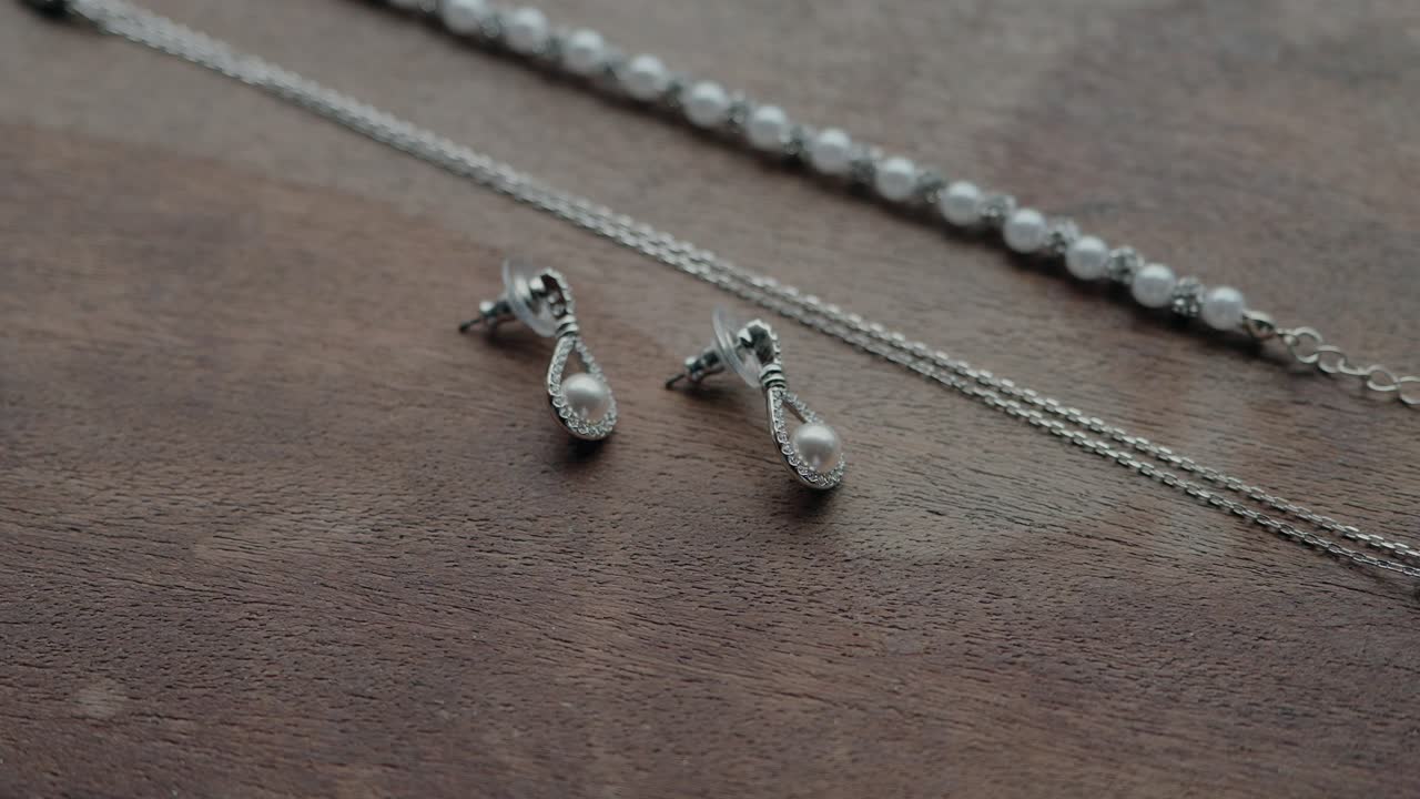 close up of pearl earrings paired with silver necklace and white beaded bracelet on wood surface