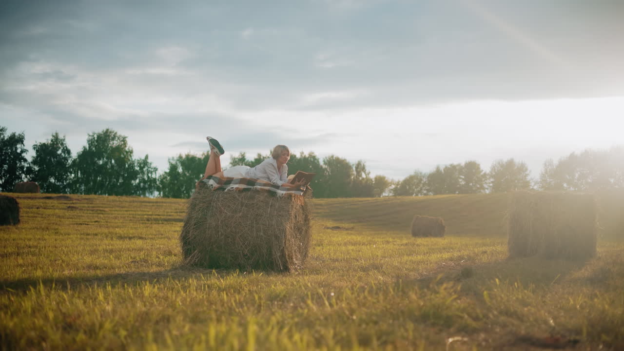 Student lying on hay bale with checkered blanket, reading book in peaceful open field, glowing sunlight effect as she flips to a new page, enjoying tranquility in nature