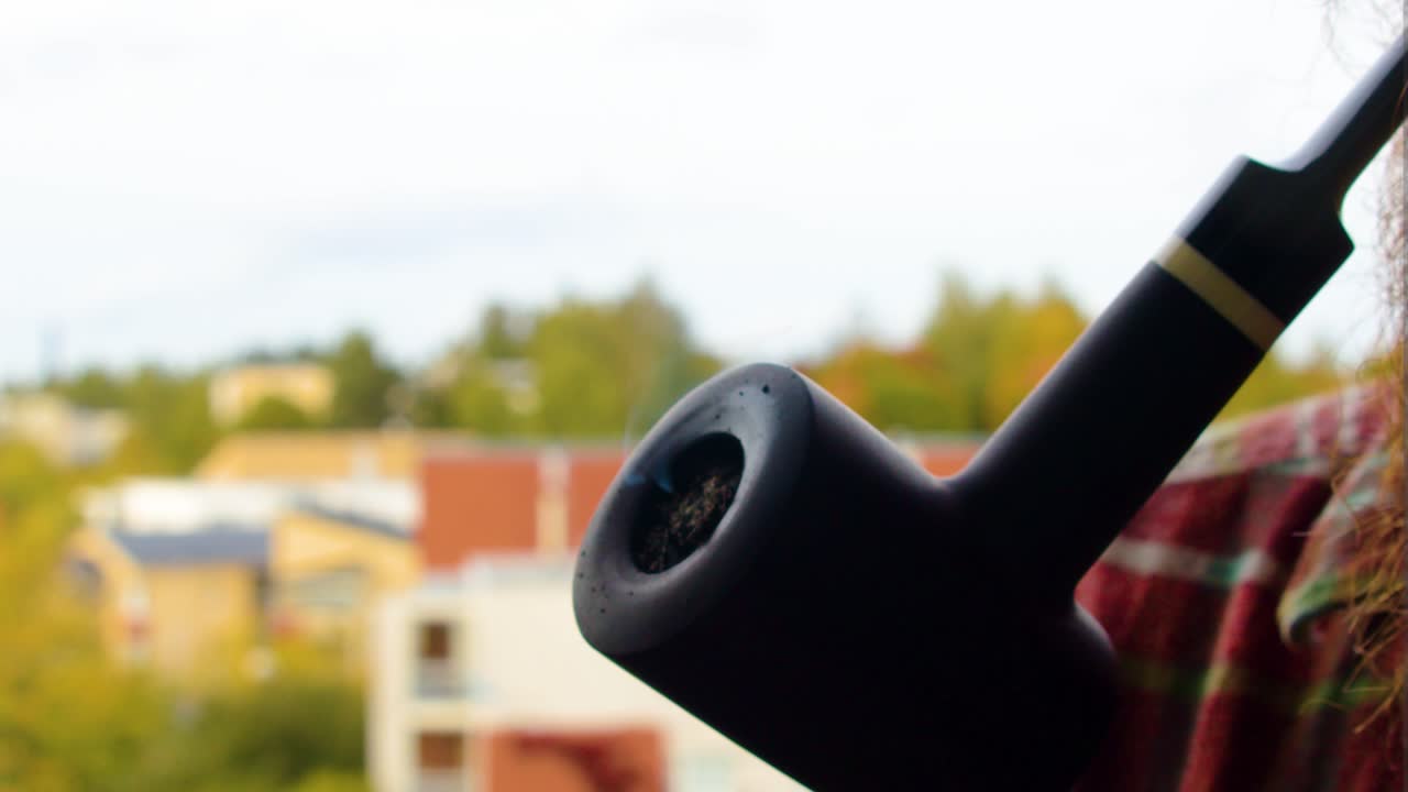 황금색 줄무 ⁇ 가 있는 검은색 빈티지 파이프의 클로즈업 흡연 담배를 발코니에서  ⁇ 은색 수염을 가진 남성이 가끔씩 들고 부분적으로 구름진 날 집을 지원합니다. 배경에는 마을이 있습니다.