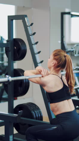 athletic woman in black sporty leggings with a ponytail is crouching with a barbell in front of a mirror in a fitness club near professional sports equipment. vertical video