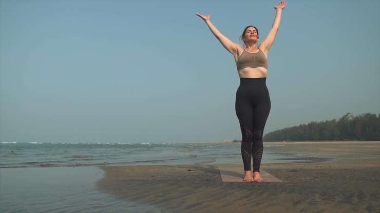 Donna in piedi che fa stretching yoga sulla riva di una spiaggia.