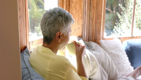 Happy-senior-biracial-woman-sitting-on-couch-and-drinking-tea-at-home,-slow-motion