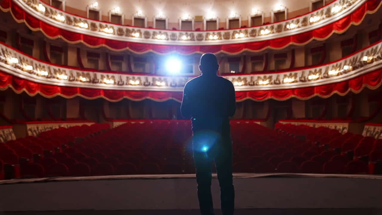 Man in front of empty theatre hall with red velvet chairs. Actor is standing on stage and talking with gestures before the performance.