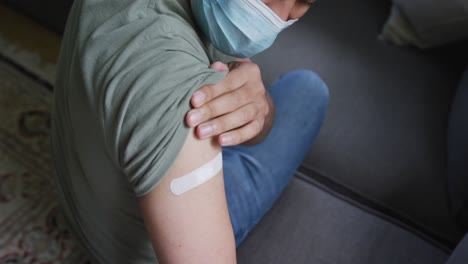 young man in protective face mask sitting on sofa after vaccination during coronavirus outbreak