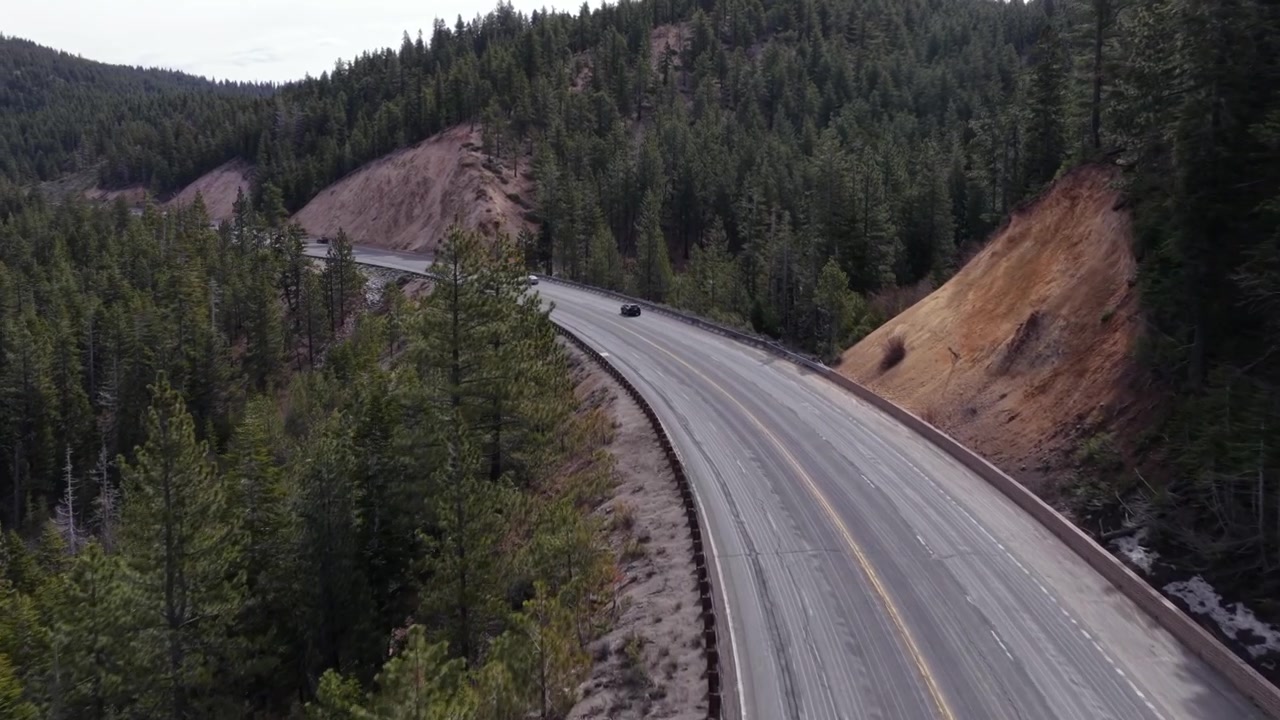 Autostrada di montagna del Nevada che si snoda attraverso una fitta pineta con tagli di collina rocciosi esposti, traffico scarso e cielo grigio coperto nella selvaggia natura del Pacifico nord-occidentale. Ripresa aerea di inseguimento