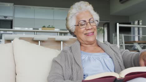 Mujer-Mayor-Afroamericana-Leyendo-Un-Libro-Mientras-Está-Sentada-En-El-Sofá-De-Casa