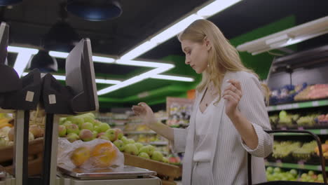 una chica rubia en un supermercado pesa naranjas en una balanza electrónica presionando la pantalla de pie con una canasta en sus manos