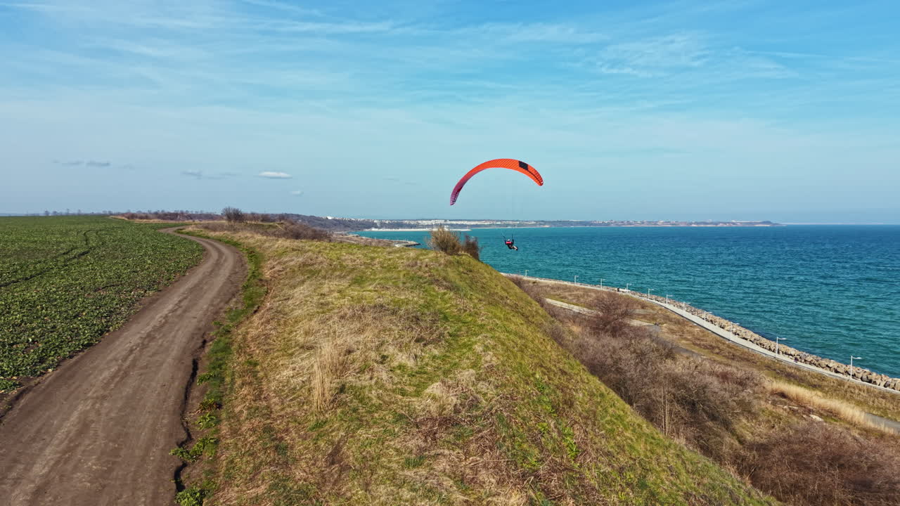 Paraglider flies over coastal field and water on a clear day near a scenic path by the shore