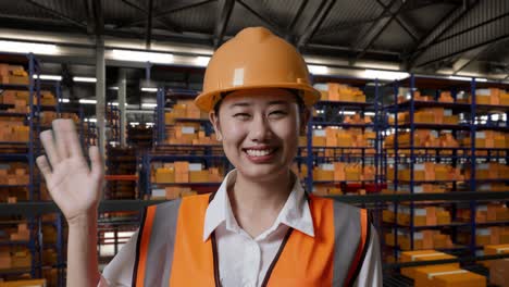primer plano de una ingeniera asiática con casco de seguridad de pie en el almacén con estantes llenos de mercancías de entrega. sonriendo a la cámara y agitando la mano diciendo adiós en el almacenamiento