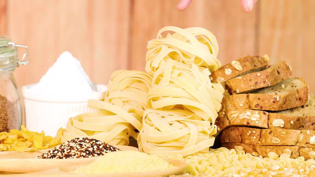 A hand selects tagliatelle pasta from a display of bread and grains in a bright, warm setting