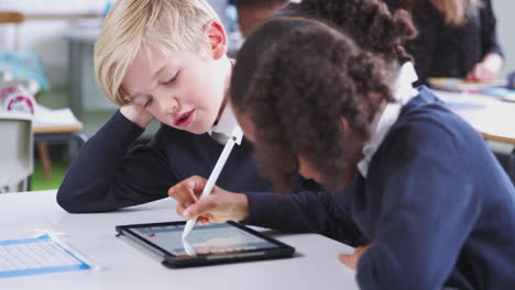 Girl-using-a-tablet-and-stylus-sitting-with-a-boy-at-a-desk-in-a-primary-school-class,-rack-focus