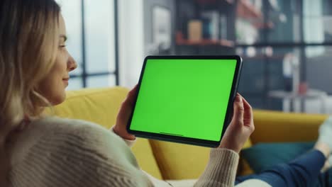 beautiful authentic female using tablet computer with green screen mock up display at home living room while lying on a couch sofa. she's browsing the internet and checking videos on social networks.