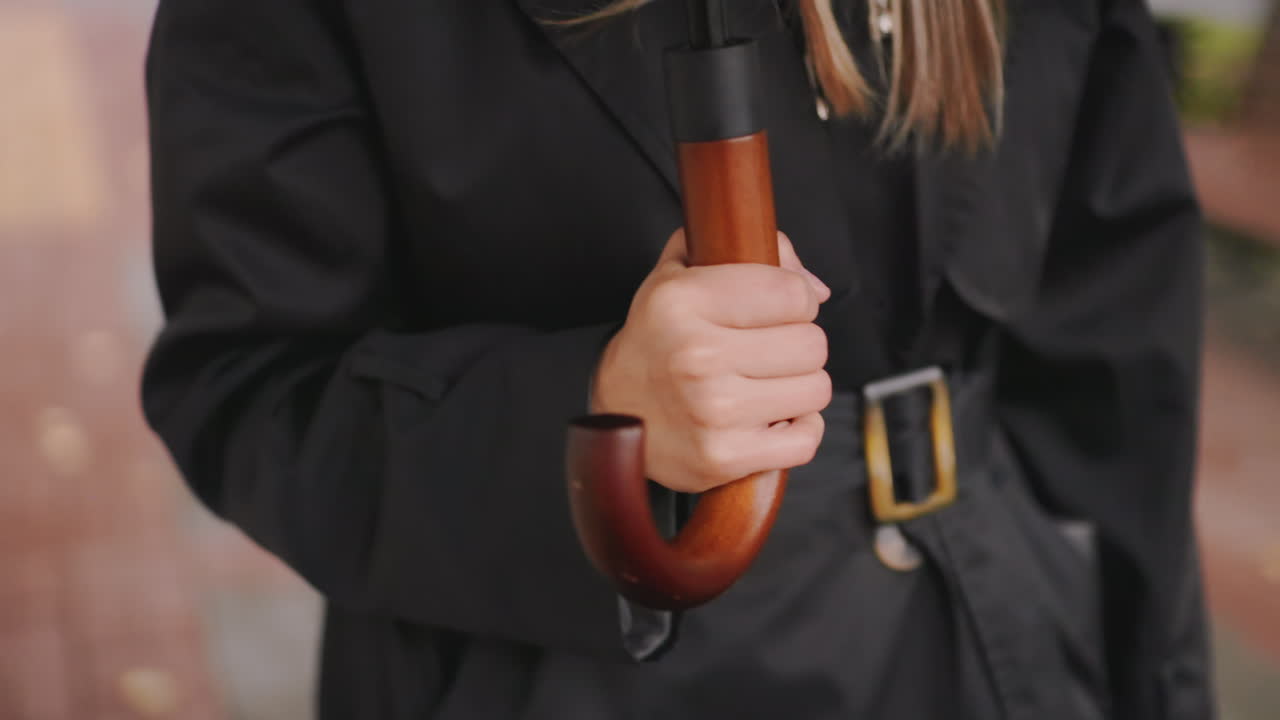 Close-up of woman hand holding wooden handle umbrella while wearing black coat with belt on rainy autumn day, blurred pavement and fallen leaves in background, detail of stylish accessory fashion