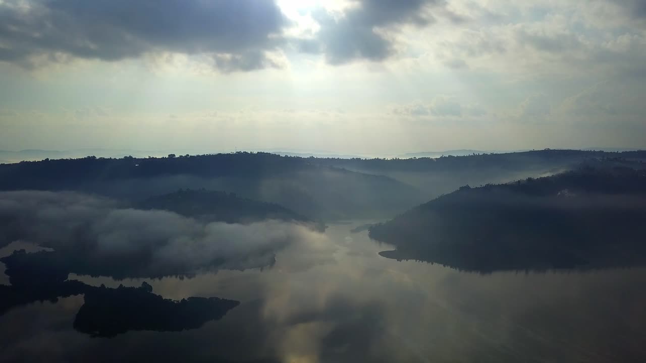 Aerial wide drone view of Lake Bunyonyi in southwestern Uganda, with mist and low lying clouds drifting over the water and terraced hills, illuminated by soft sunlight