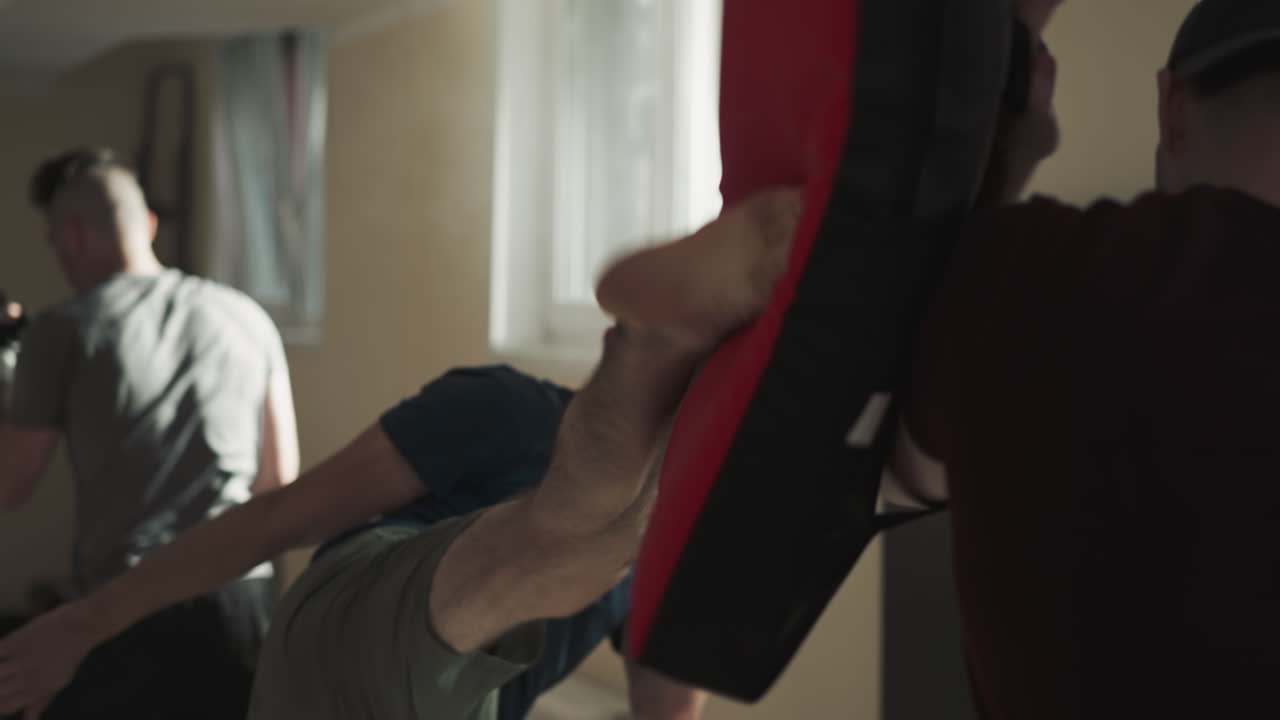 튼튼한 젊은 남자 권투 선수는 특수 장비와 싱백을 사용하여 강한 다리 타격 기법을 보여줍니다. 어두운 체육관에서 그룹 훈련 세션. 참가자에게 트레이너의 지침