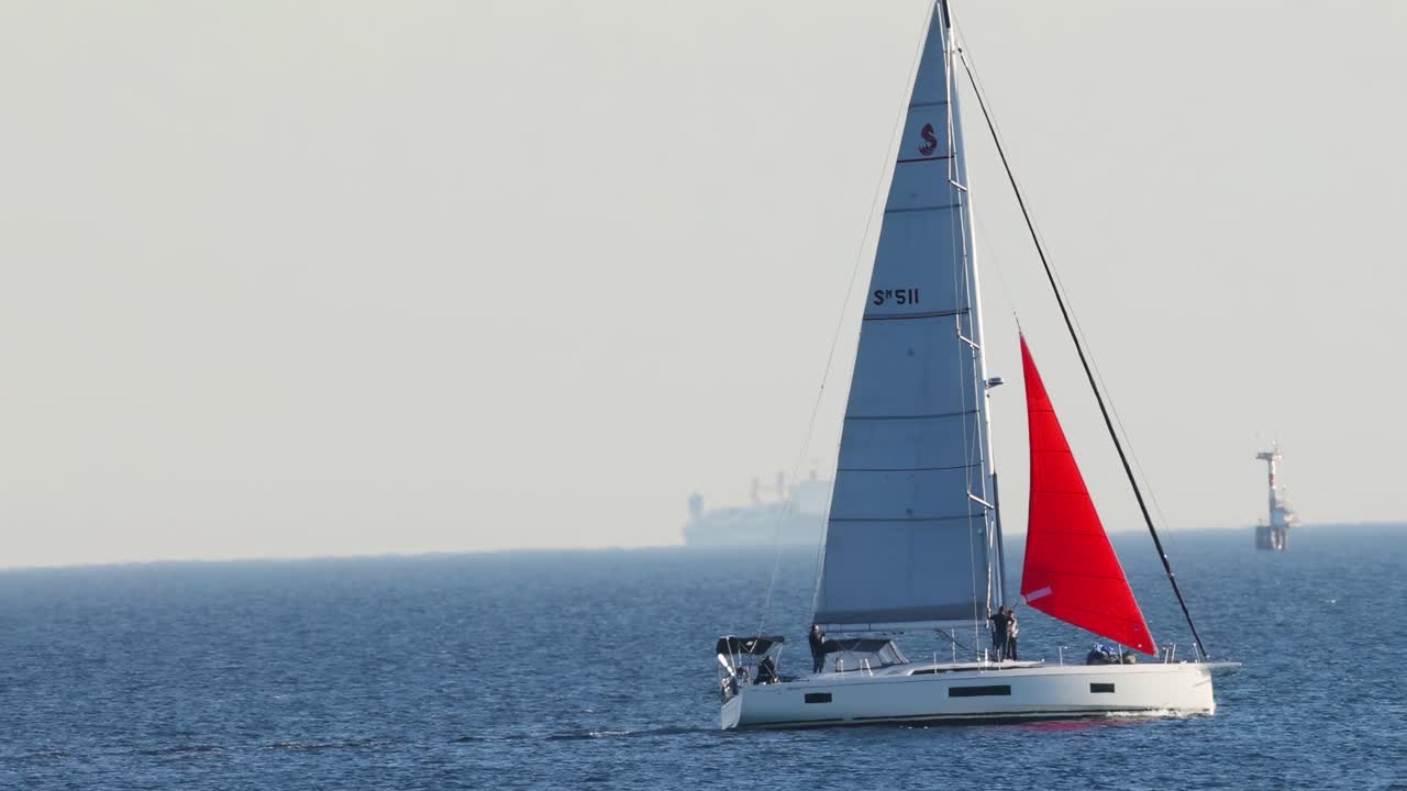 A sailboat with a striking red sail navigates calm blue waters under a clear sky.