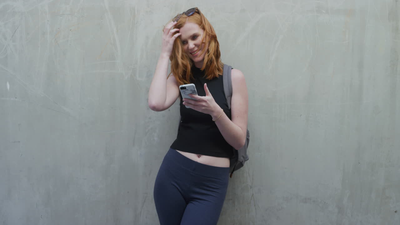 ritratto di una bella giovane donna dai capelli rossi che usa uno smartphone che si gode di navigare messaggi di testo su uno sfondo di muro di cemento del telefono cellulare