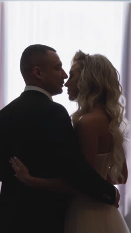 young man silhouette in elegant suit kisses nose of blonde curly haired woman standing together against window at honeymoon backside view slow motion