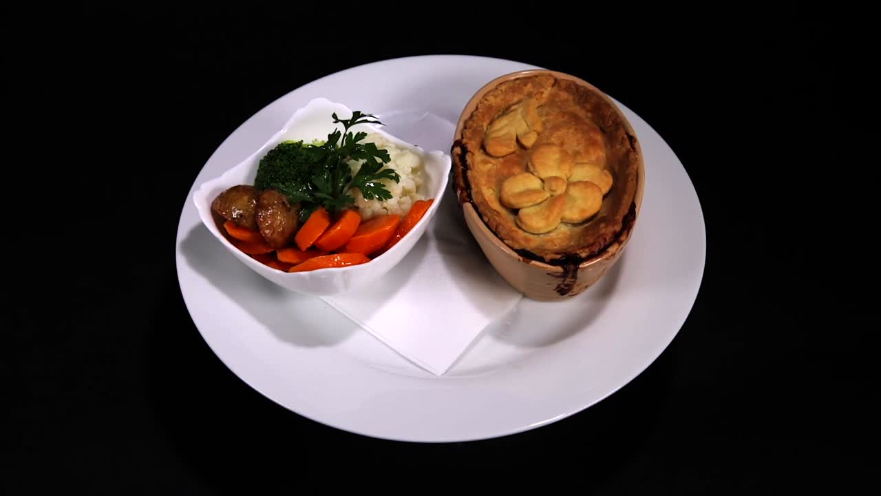 Spinning plate with golden meat pie and colourful steamed vegetables, black background.