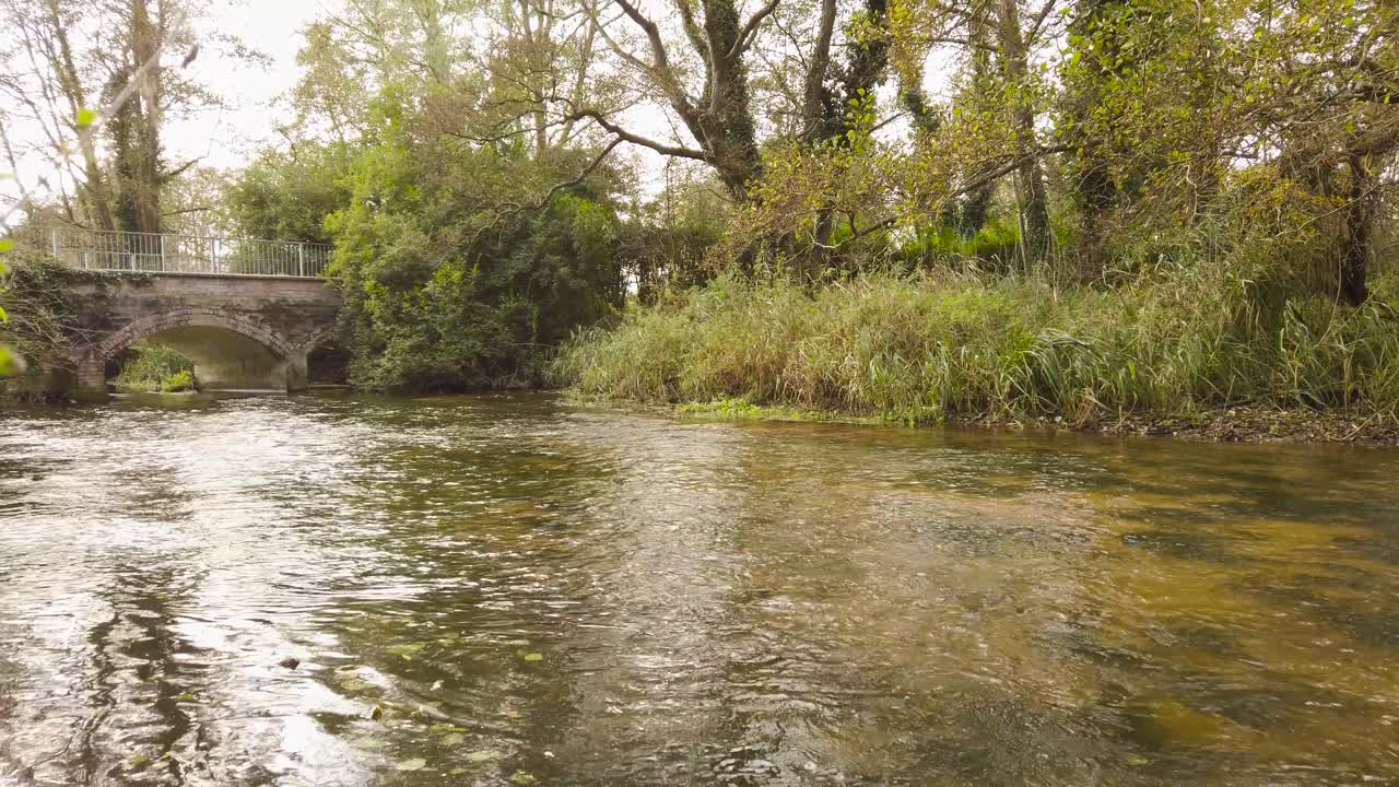 An abstract dolly shot revealing the beautiful shallow waters of the flowing Thetford Little River, in the distance an old arched bridge crossing over the river in Norfolk, England