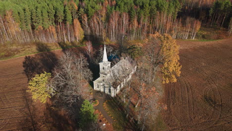 Drone-shot,-Birds-eye-view-of-old,-charming-countryside-church-surrounded-by-fields-and-forest-in-autumn