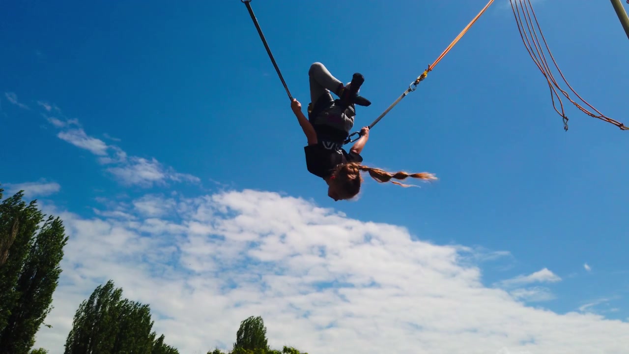 Ripresa statica dal basso cattura un bambino imbracato in un sistema elastico di bungee, che esegue capriole in avanti e all'indietro in alto nell'aria contro un cielo azzurro brillante a un festival primaverile nel parco.