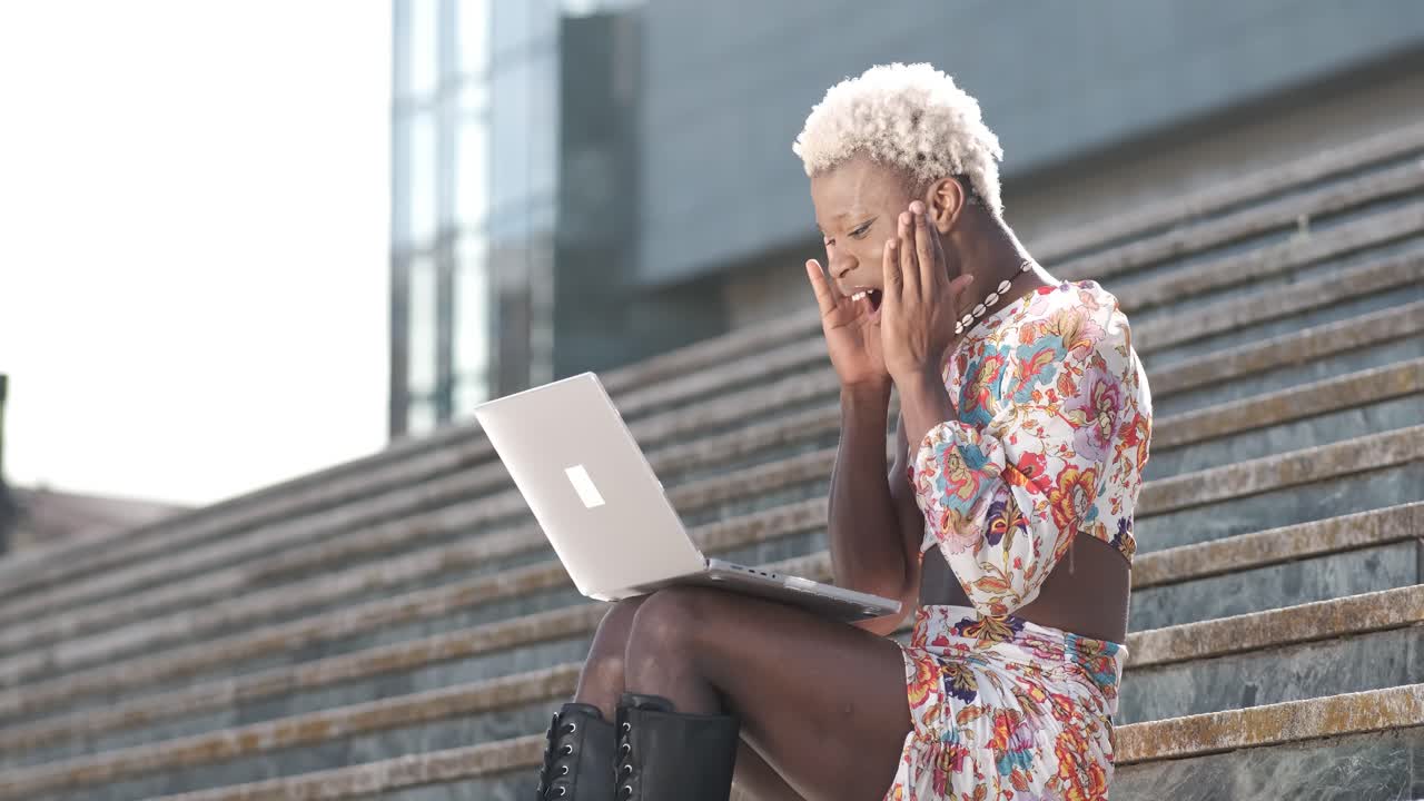 Transgender person looking surprised and celebrating while using a laptop sitting on the stairs outdoors.
