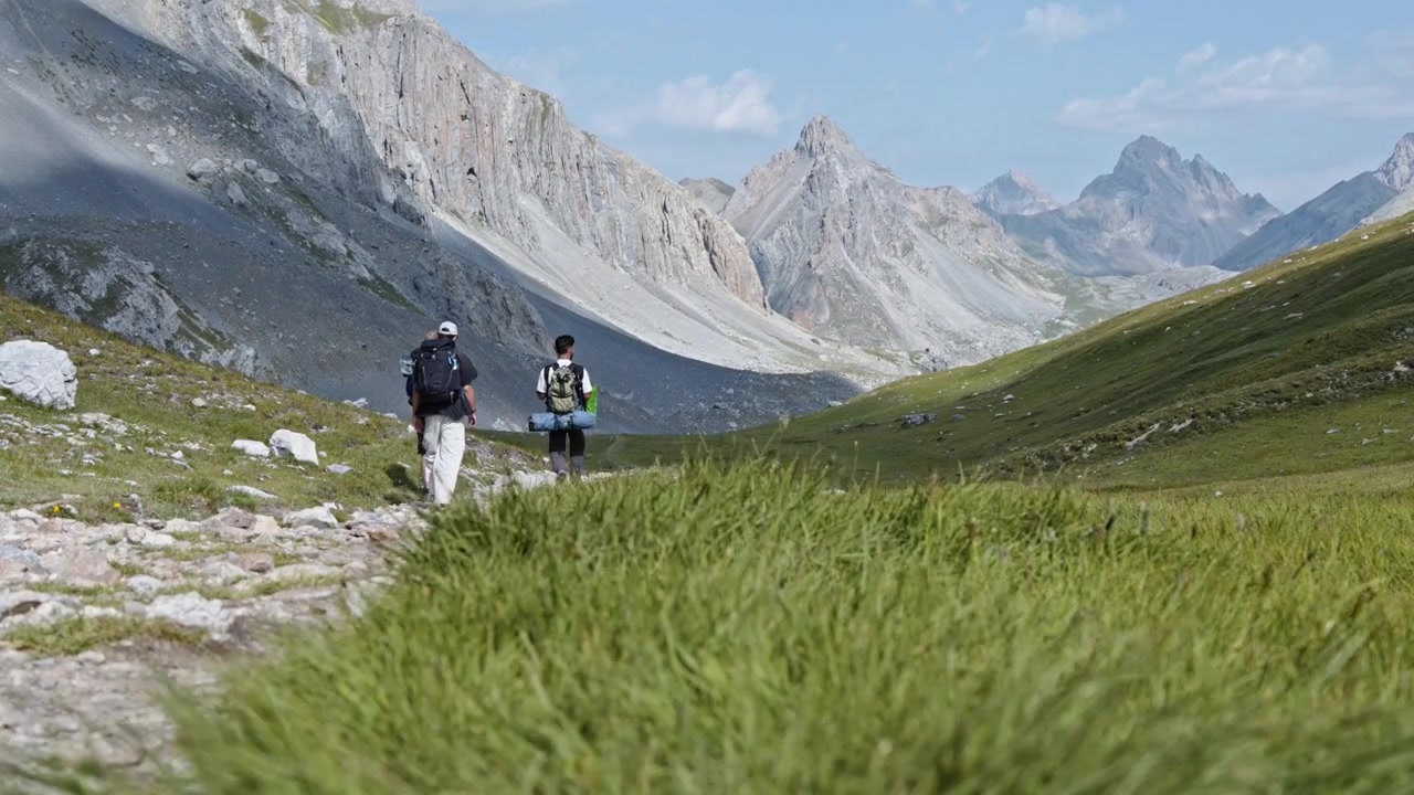 Tre viaggiatori con zaino in spalla camminano lungo un sentiero roccioso attraverso un'ampia valle alpina verde incorniciata da ripide cime italiane in una limpida giornata estiva