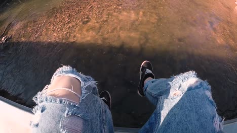 ultra slowmotion en primera persona punto de vista de las piernas de un joven con vaqueros rasgados balanceándose mientras se sienta en el borde de un puente sobre un río que fluye durante un día soleado