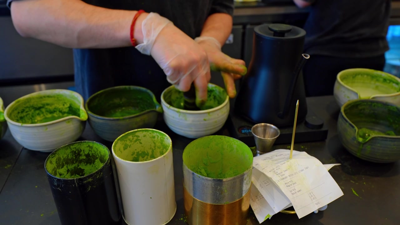 Barista preparing matcha tea in ceramic bowls at cafe counter. Gloved hands scooping green powder among bowls and metal canisters.