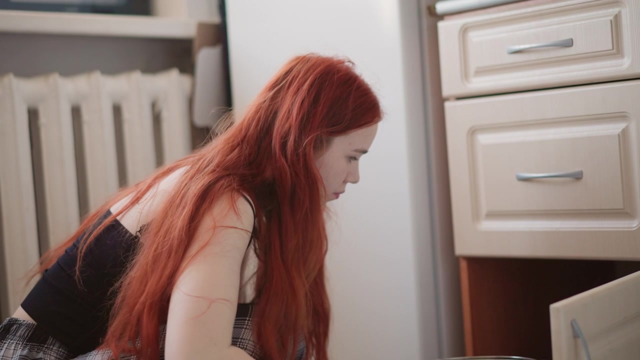 Mulher ruiva branca ajoelhada ao lado do armário, abrindo a gaveta e pegando uma caixa, ação de recuperação focada, busca em armazenamento inferior da cozinha, momento prático de organização doméstica, expressão intencional.
