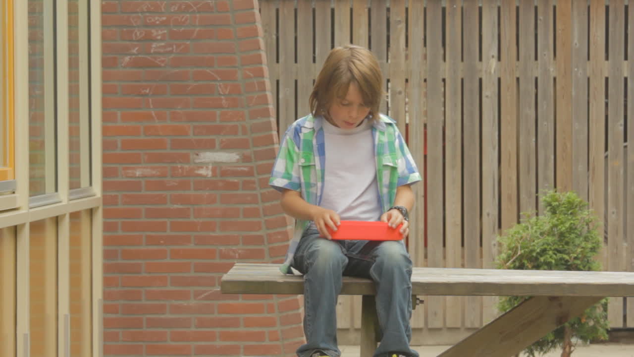 Un giovane ragazzo solitario che mangia il suo pranzo su un tavolo nel cortile di una scuola con bambini che corrono intorno.