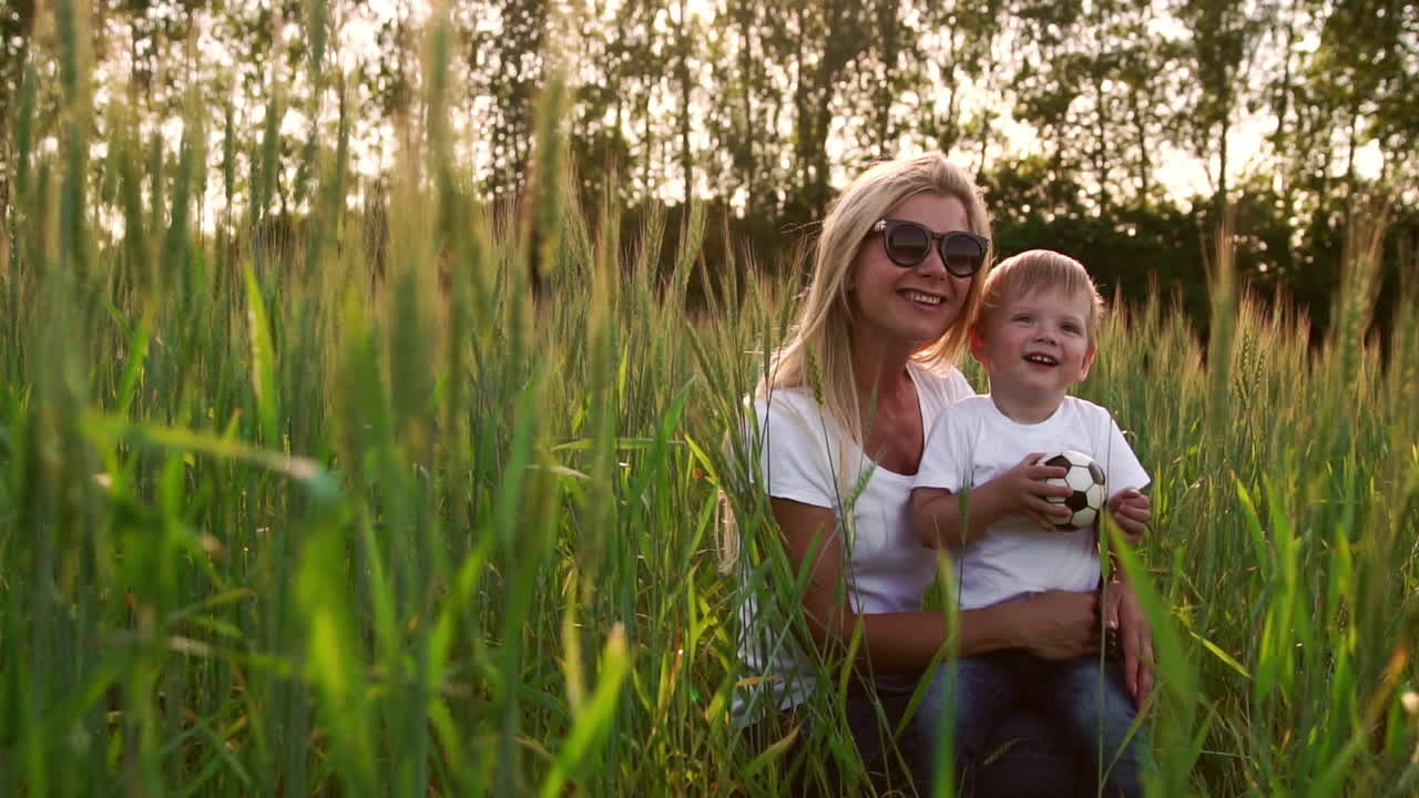 amorosa madre e hijo abrazándose y jugando con una pelota de fútbol en un campo con espiguillas a la hermosa luz del atardecer con camisetas blancas