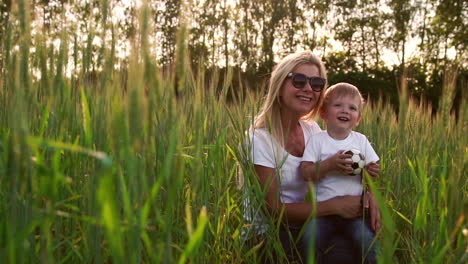 Loving-mom-and-son-hugging-and-playing-with-a-soccer-ball-in-a-field-with-spikelets-in-beautiful-sunset-light-in-white-t-shirts