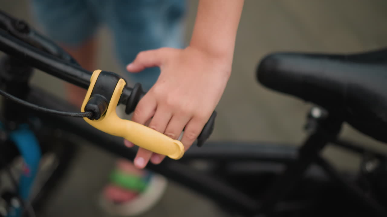 un primer plano de la mano de un niño agarrando el freno de una bicicleta, el enfoque está en la mano del niño y la palanca de freno amarilla, mientras que el fondo permanece borroso