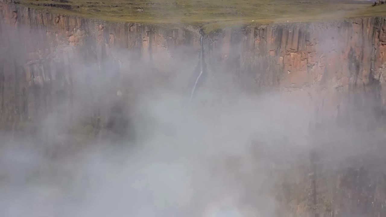 Aerial drone footage of Tugela Falls waterfall narrow river water falling down a steep brown tall cliff creating steam and mist in the sunny South Africa air. Cliff has green plants growing on it.