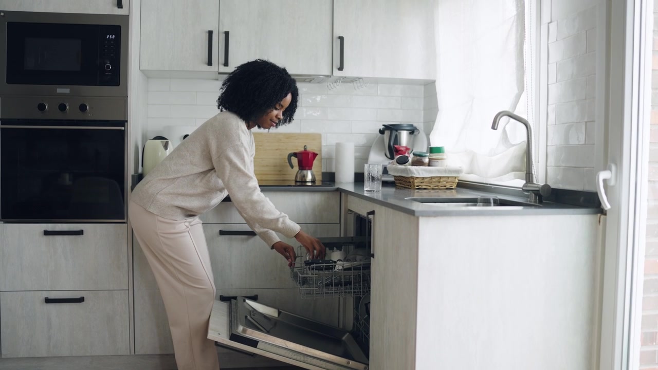 Giovane donna afroamericana che carica i bicchieri nella lavastoviglie in una cucina moderna.