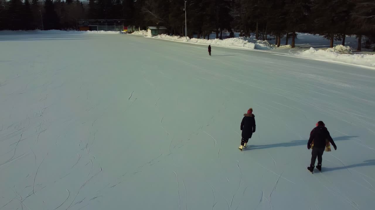 sobrevuelo aéreo de 4k detrás de parejas que tienen citas dobles en una pista de patinaje sobre hielo hecha por el hombre rodeada de árboles altos junto a un campo de golf del valle cubierto de nieve por condominios de torres residenciales con vistas a ellos