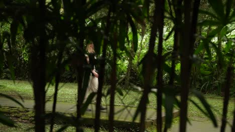 Attractive-young-female-explorer-going-through-the-jungle-on-a-paved-stone-path-shot-through-the-bamboo-tree-in-a-shade-of-rain-forest-with-backpack-walking-confidently-Ubud-Bali-Indonesia-slow-motion