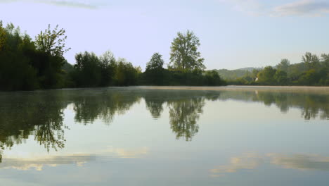 una vista de la superficie del lago al amanecer cuando la niebla se eleva sobre el agua y el sol brilla detrás de un árbol que se arrastra sobre el lago con un montón de nubes reflejadas en el agua