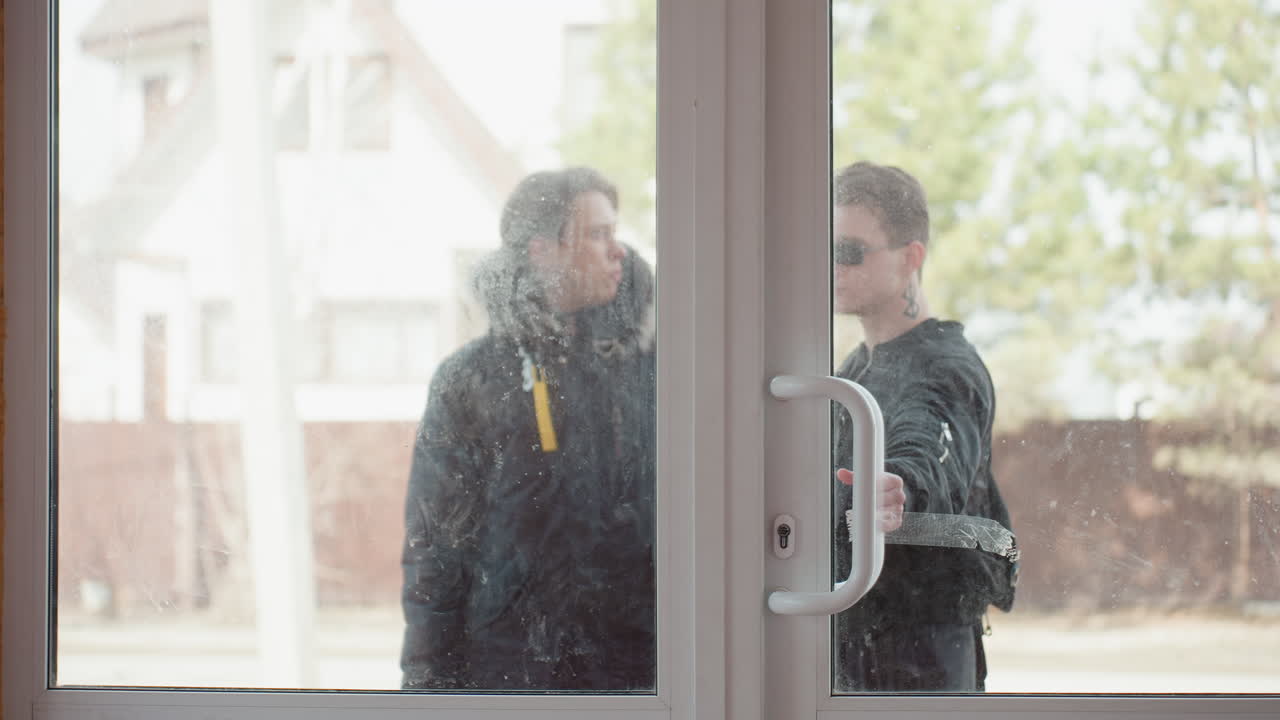 Two guys in dark clothing walking into a gym , slightly blurred through dirty window, one wearing sunglasses, shapes and movements visible behind handle, sunlight reflecting off glass, suburban background