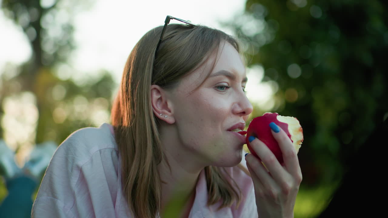 cerca de la joven que ríe suavemente mientras toma un bocado de la manzana roja en su mano, las piernas cruzadas en la rodilla, con un hermoso fondo borroso bokeh y la cálida luz del sol filtrando a través de los árboles