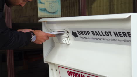 young man drops mail-in ballot letter in slot at official voting box with drop ballot here sign