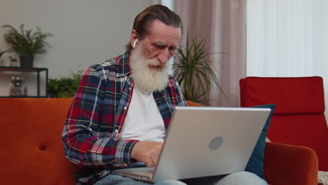 senior grandfather man freelancer working on laptop, sends messages, makes online purchases at home
