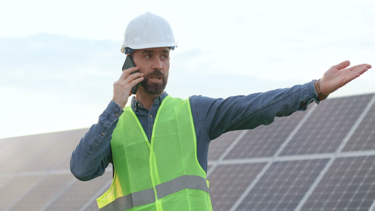 hombre caucásico con uniforme especial y casco protector y dando instrucciones y hablando por teléfono en una plantación solar