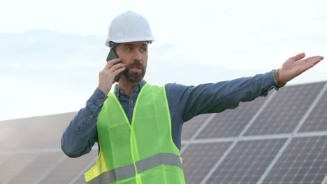 caucasian man in special uniform and protective helmet and giving instructions and talking on the phone on solar plantation