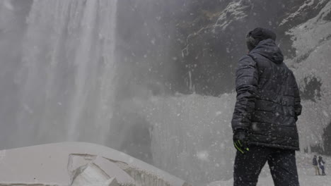 Back-of-Man-in-Warm-Clothes-Walking-on-Snowfall-Under-Waterfall-in-Cold-Winter-Landscape-of-Iceland