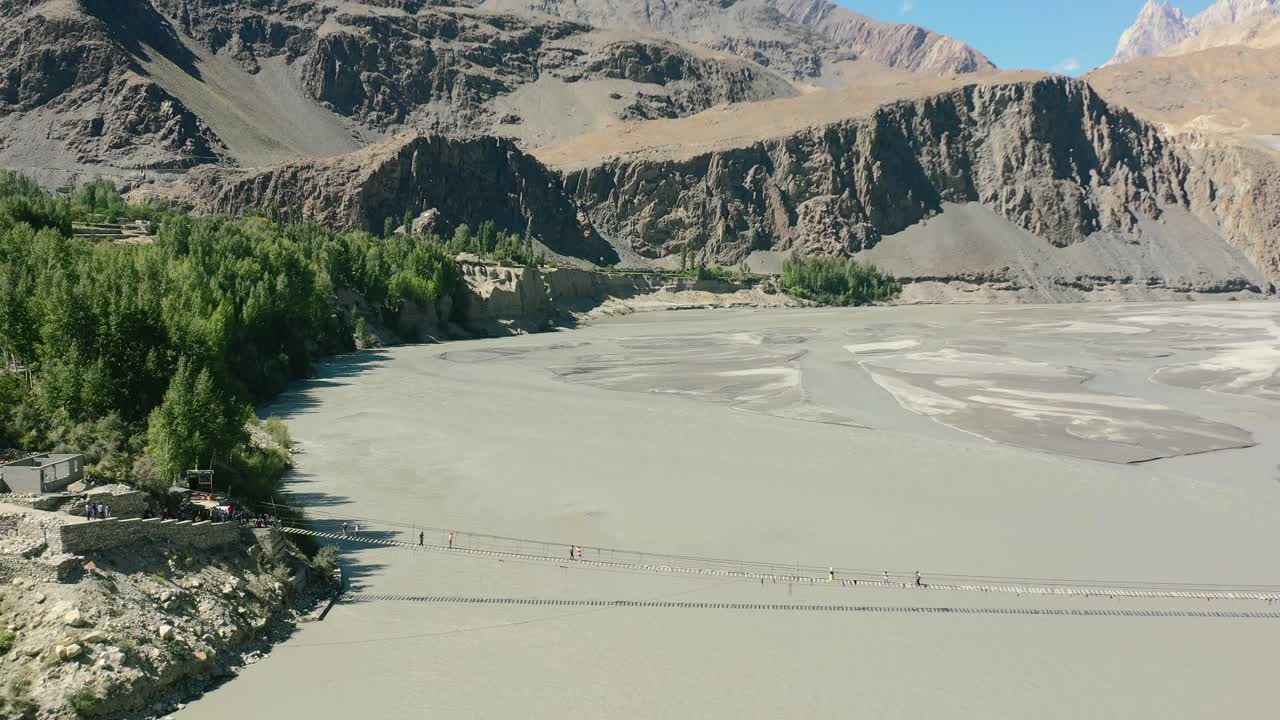 drone aéreo volando hacia atrás mientras los turistas cruzan el famoso puente hussaini en hunza pakistán con un río rápido que fluye debajo y un bosque verde bordeando el río