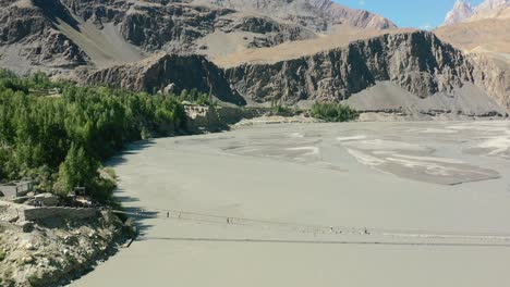 aerial drone flying backwards as tourists cross the famous hussaini bridge in hunza pakistan with a fast river flowing below and a green forest bordering the river